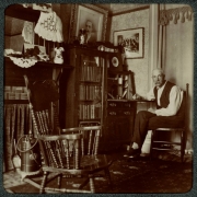 A man sits at a fold-out desk and bookcase in a home in Denver, Colorado. A rocking chair and fireplace are in the room. Photographs hang on the wall.