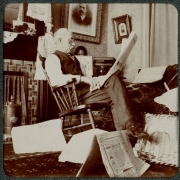 A man sits in a rocking chair and reads a newspaper in his home in Denver, Colorado. A fireplace is in the room and photographs and portaits hang on the walls. Newspapers litter the floor.