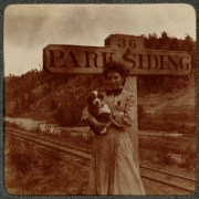 A woman poses with a puppy near a sign that reads: "36 Park Siding" on the Colorado and Southern Railway (C&S) route at Park Siding, (Jefferson County), Colorado. Railroad tracks are nearby.