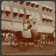 A man and woman ride in an automobile, as part of a parade, on 17th (Seventeenth) Street in Denver, Colorado. Spectators stand on the sidewalk and look on from windows in the Drexel Hotel. Signs read: "[?]ssed Brick", "Fire Brick", Drexel Ho[tel]", and "Welsbach Lamps".