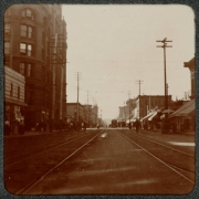 View, facing northwest, of the Mining Exchange Building and 15th (Fifteenth) Street from Curtis Street in Denver, Colorado. Pedestrians walk on and near the street. Trolley tracks are in the center of the street. A sign reads: "Sample Store".