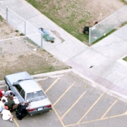 A student lies on a sidewalk while an officer guards others behind a car.
