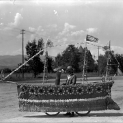 A man stands in a bicycle float shaped like a steam ship, Sunflower Carnival parade, Colorado Springs, El Paso County, Colorado. The float is decorated with paper and flowers. One of the masts bears a flag that reads: "Colorado". A U. S. flag flies from another mast. Photo taken in August.