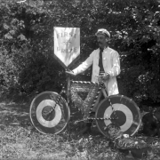 A man poses near a group of trees with a safety bike and a banner that reads: "First Prize". The bike is decorated with flowers, bunting and streamers. Two dogs sit near the bicycle. Photo taken in August