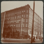 View of the Charles Building on the corner of 15th (Fifteenth) and Curtis Streets in Denver, Colorado. Pedestrians walk on the sidewalks and in the street near horse-drawn buggies. A sign reads: "Chickering Pianos".
