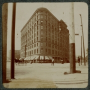 View of the Brown Palace Hotel on the corner of Broadway and Tremont Place in Denver, Colorado. Pedestrians, a bicyclist, and a man in a horse-drawn buggy cross Broadway near the building.