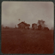 Men walk near a building and water tank in a train yard, possibly in Denver, Colorado. A caboose is on the tracks nearby.