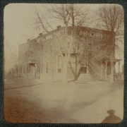 View of the Hotel Brighton (formerly the Platte Valley House) located on the corner of Bush and Cabbage Avenue in Brighton (Adams County), Colorado. Shows a two story brick building with porches, a balcony, and chimneys. Signs read: "Hotel Brighton."