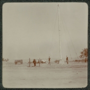 Men in military uniforms walk near a flagpole possibly in Denver, Colorado. Cannons and wooden wagons are nearby. The flagpole is tethered by guy wires.
