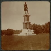 Women pose on the base of a statue of Robert Burns in City Park, Denver, Colorado. The women wear long dresses and hats decorated with flowers. The base of the statue reads: "Burns".