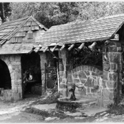 View of the spring house at the upper switch back of the Lariat Loop Road on the way to Lookout Mountain Park, a Denver Mountain Park located 17 miles west of Denver, Colorado. Shows two rock ashlar buildings and a water pump in front of a arched rock wall. The shelter has an arch window, a stone bench and shingled roof.