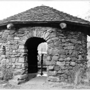A view of a pump house at Little Park, Jefferson County, located 21 miles southwest of Denver, Colorado. The round shelter has rock ashlar walls, two arched doorways and a shingled pyramidal roof. The pump is in the center of the shelter.