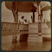 Women pose in the floating bandstand in City Park, Denver, Colorado. They wear long dresses and hats. A wooden bridge connects the bandstand to the rest of the park.