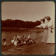 Women feed geese near Ferril Lake in City Park, Denver, Colorado. They wear long dresses and hats decorated with flowers.