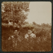 Women pose behind a bush in City Park, Denver, Colorado. They wear hats decorated with flowers.