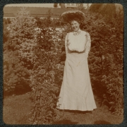 A woman poses outdoors near a shrub at her home in Denver, Colorado. She wears a long dress and a hat decorated with flowers.