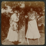 Women pose outdoors near a tree near thier home in Denver, Colorado. The wear long dresses and hats decorated with flowers. One woman holds a handbag.