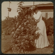A woman poses outdoors near a shrub at her home in Denver, Colorado. She wears a long dress and a hat decorated with flowers.