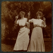 Women hold drinks in glass bottles and, probably, fly a kite in their yard in Denver, Colorado. They wear long dresses.