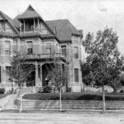 A man, probably Amos Steck, stands on the porch of a house at 1308 Glenarm Place in Denver, Colorado. The three-story brick masonry house has turrets, covered porch, and balconies. Summer, 1886.