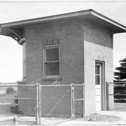 Shows the small brick lighting control building at the Denver Municipal Airport (later Stapleton International Airport) in Denver, Colorado. The building features a single door, windows and a flat roof with an overhang; a chain link fence is in front of the building.