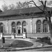 Shows a view of the Roger W. Woodbury branch library in Denver, Colorado located on West Highland Place and Federal Boulevard. The one-story Renaissance style building was designed by J.B. Benedict and features red tiled roof, arched windows and doorways and a front balustrade. The Woodbury Branch was opened in 1913.