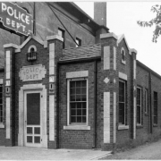 An exterior view of Police Station number 4 located on West 26th Avenue and Federal Boulevard in Denver, Colorado. The single story brick building features cement trim and accents and a shingle roof. A sign above the entrance reads: "Police Dept."; and a neon sign above the building reads: "Police Dep't".