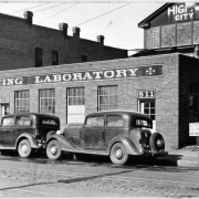 Automobiles are parked in front of the single story brick testing laboratory of the Denver Highway Department located near 8th (eighth) and Walnut Street in Denver, Colorado. Address above two doors of the building read: "811" and "805". Garage number one is in the background.