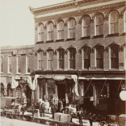 View of Larimer Street businesses in Denver, Colorado; shows brick storefronts, men posing, merchandise, a horse-drawn delivery wagon, and signs: "Daniels & Fisher Co," "J. H. Hense & P. Gottes," "Jewelry Manufacturers," and "Miller's Boston Boot and Shoe Store."
