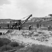 Denver and Rio Grande Western Railroad construction in Ruby Canyon, (probably) Utah consists of men laying track over a concrete bridge, using a rail-adapted crane. Sandstone cliffs are in the background.