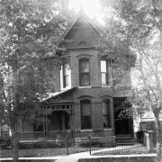View of a two story house designed and built by Orlando Scoby on 2743 Curtis Street in the Curtis Park Historic Districts in Denver, Colorado. The brick house has a porch with decorative wood trim, a pitched roof, and iron fence. A flagstone carriage stoop is in front of the house.