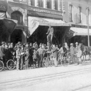 A large group of men are gathered around bicycles, one of which has a seat ten or more feet in the air, Denver, Colorado. Signs read: "G. E. Hannan Bicycles," and "Adams Express Company."