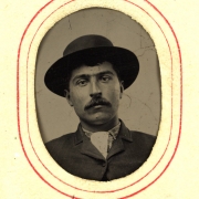 Studio portrait of Hammer. He has a moustache and wears a coat, scarf tie, and wide-brimmed hat.
