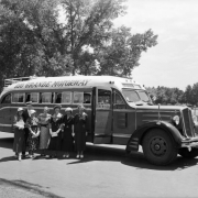 View of a Rio Grande Motor Way passenger bus. Women and a man pose by the 1935 White brand vehicle; outfits include print dresses and straw hats. Pin striping reads: "Los Angeles - Salt Lake - Grand Junction - Pueblo - Denver - Omaha - Kansas City - Chicago - New York 154;" possibly City Park Lake is in the background.