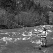 A young woman fishes with a bamboo pole, in (probably) Colorado or Utah; she wears hip waders and stands in the river. Her outfit also includes short-legged bib-overalls and a straw hat.