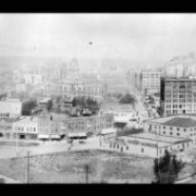 Panoramic view of Denver, Colorado looking west; landmarks include the Arapahoe County Courthouse, Fire Engine House Number One, Brown Palace Hotel, the Majestic Building, and Hotel Metropole. Signs read: "Hair Dressing," "The "M" Company of America," "Palace Stables," "Geo. Sell's Bakery," "A. T. Wilson. Automobiles. Repairs. Storage.," "Denver Clothes Pressing Co. $3.00 For # Months.," and "Drugs, Prescriptions, Cigars At Corner."