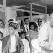 Men, women, and children stand near a building during the George Washington Carver Day Nursery dedication at 2260 Humboldt Street in the City Park West neighborhood of Denver, Colorado. Burnis McCloud stands by a camera near the group.