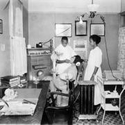 Clarence F. Holmes, an African American man, works on a patient while his assistant stands nearby in his office at 2602 Welton Street in the Five Points neighborhood of Denver, Colorado. Dentists' tools, a desk, medicine chest, and a sink are in the room.