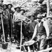 A group of miners pose standing near a wheelbarrow, Yukon Territory. One man holds a pan full of placer ore, the others hold shovels; mining construction is behind them.