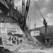 View of the Colorado Yule Marble Company quarry, four miles south of Marble (Gunnison County), Colorado; shows a crane, buildings, cable and pulleys, a suspended block of stone, a gondola, and men working. The block will be transferred to the cable the gondola is on for lowering to the standard gauge electric railway for transport to the mill site, in the town of Marble.