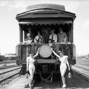 Men and women pose on the observation platform of the "Glenwood Canon" Denver and Rio Grande Western Railroad passenger coach at Burnham Shops, Denver, Colorado. Ladies in bathing suits and conductor's hats pose with lanterns.