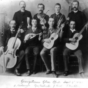 The Georgetown Glee Club poses in front of a painted studio backdrop with their instruments, Georgetown, Colorado. Eight men wearing suits and ties form two rows. Instruments include a cello, a mandolin, guitars, violin, and a cornet. Label tapped at bottom of original identifies some men as: "P Ashcroft, Geo Rubado, J. Keek, T. Rodda."