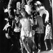A young Mexican American boy in an Aztec dance costume dances. He wears a smock with decorative trim, a headdress with feathers and he holds an arch of paper flowers.