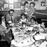 Interior view of the Ringside Lounge at 1120 17th Street in Denver, Colorado; shows boys, girls, a man and a woman at a table set with food and dinnerware. Joe "Awful" Coffee offers a piece of cake; Jane Sterling holds a child, and framed photographs cover the wall.