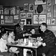 Interior view of the Ringside Lounge in Denver, Colorado; shows boy boxers, Joe "Awful" Coffee shaking hands with Father Jim Moynihan, and a table set with food. Framed pictures of boxers and newspaper clippings cover the wall.
