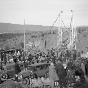 People crowd the Royal Gorge Bridge for its dedication in Fremont County, Colorado. A woman in a fur coat is by automobiles; United States flags and bunting decorate a dais.