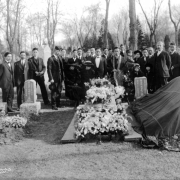 Shows a group of Japanese-American men, women and children at the funeral of James Saburo Takumi  in Denver, Colorado. A minister is in front of a flower covered casket. Two women dressed in black and with hats are seated with their heads bowed, possibly widow and family member. Tombstones with Japanese characters carved into them are in front of the group.