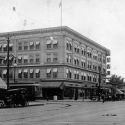 View of the intersection of 1st (First) Avenue and Broadway Street in Denver, Colorado; shows the 1st Avenue Hotel, the Citizens Exchange Bank, parked automobiles, a Denver Tramway Corporation street car, and tracks.