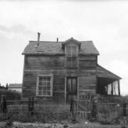View of a large, dilapidated house in Gold Hill, Colorado; features a side gable, gabled dormer, an attached, closed porch, single leaf door, double hung windows, and a horizontal timber fence with a picket gate that surrounds the property; tall grass in the yard and surrounding area.