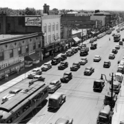 Rooftop view of Broadway at 1st (First) Avenue in Denver, Colorado. Automobiles and streetcars are on the street. Signs on buildings read: "[W]illiams Stores Stetson Hat," "Walco's Sport Spot," "Prescriptions," "Taylors," "Bernard's Proven Better Buys Men's & Boy's WEar," "Vogue Portraits," "The Skylark," "Rex Market," "Friends Electrical Appliances," "Ervington Floral Shop," "Acme Liquor Store," "Falby Paint Glass Hardware," "Crown Furniture," "Murphy Fine Food," and "Miller's."